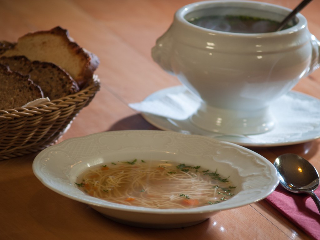 Nudelsuppe im Teller, dahinter die Suppenterrine und ein Brotkorb