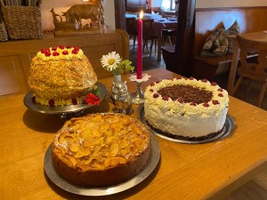 Hausgemachter Apfelkuchen und zwei Torten mit Kirschen auf einem Holztisch, daneben Kerze und Margerite